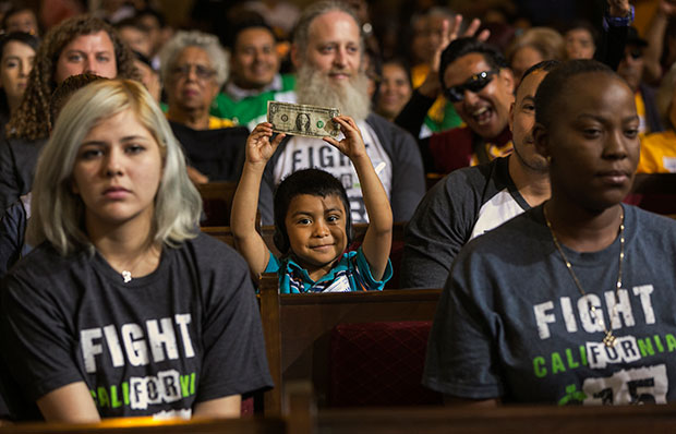 A child, center, with his father, right, raises a dollar bill as workers await the Los Angeles City Council's vote to raise the city's minimum wage to $15 per hour by 2020, June 3, 2015.